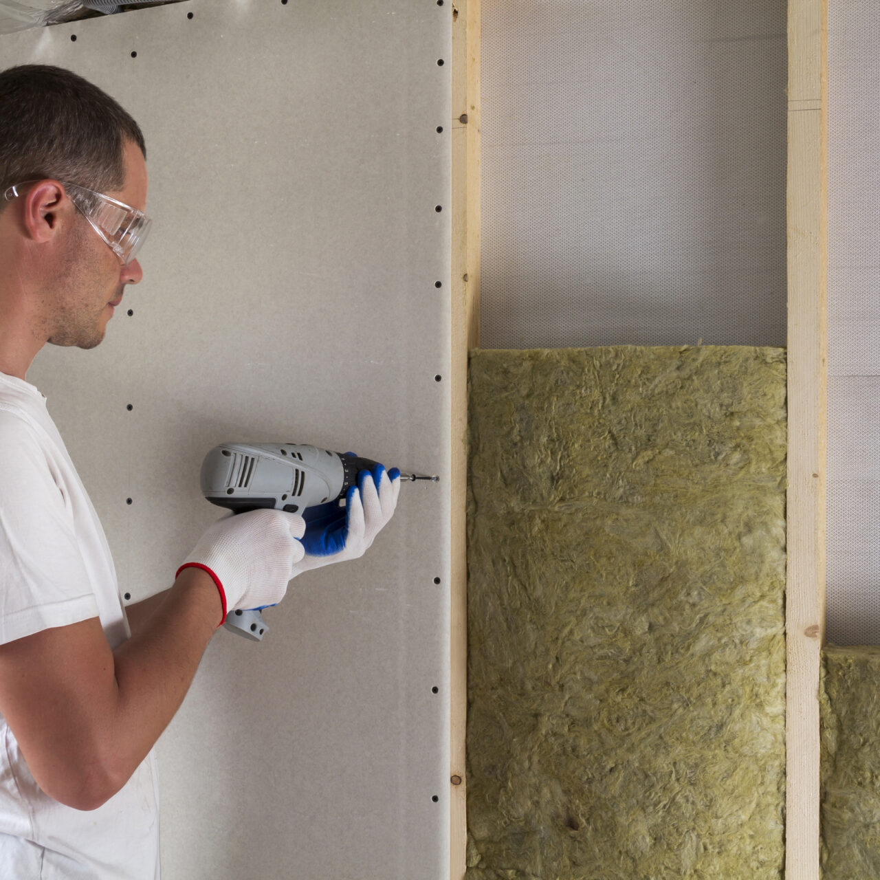 Worker in goggles with screwdriver working on insulation. Drywall on wall beams, insulating rock wool staff in wooden frame. Comfortable warm home, economy, construction and renovation concept.